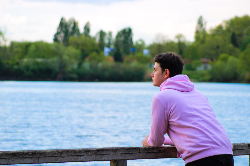 Portrait of a young man in profile in front of a lake and a forest. Teenager in pink sweater looking away on the water