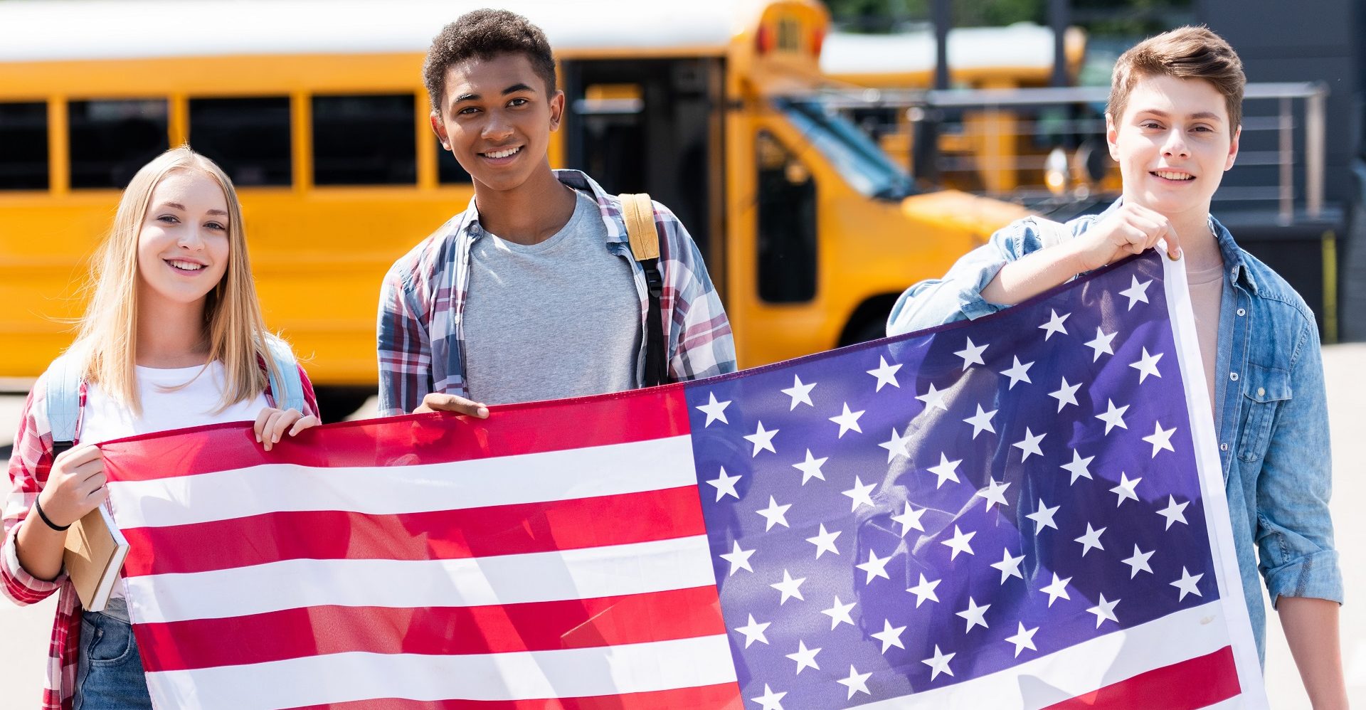 group of happy teen students holding usa flag in front of school bus
