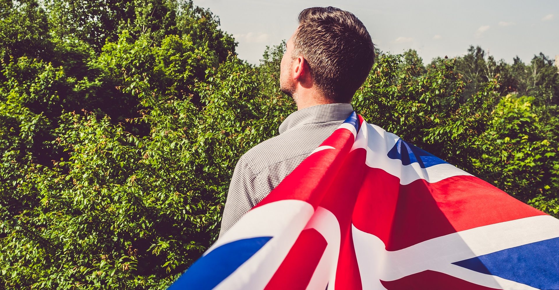 Attractive man waving a British Flag against a background of trees and blue sky. View from the back, close-up. National holiday concept